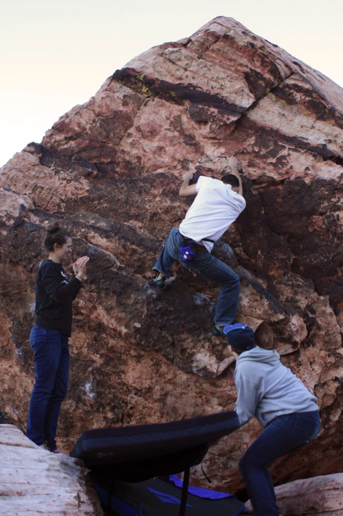 Climber going up a large boulder, spotters moving crash pads